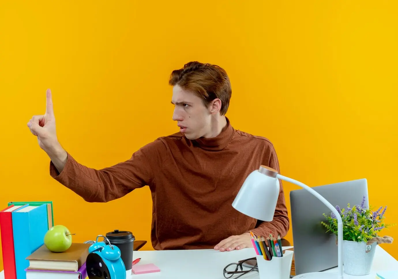 Strenger junger Schüler sitzt an einem Schreibtisch mit Schulsachen, blickt zur Seite und streckt die Hand seitlich aus.